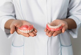 Two hands holding a complete arch of dentures each in front of white jacketed abdomen