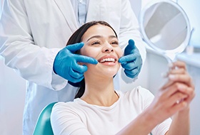 Dentist in blue gloves pointing to patient’s smile as she holds mirror